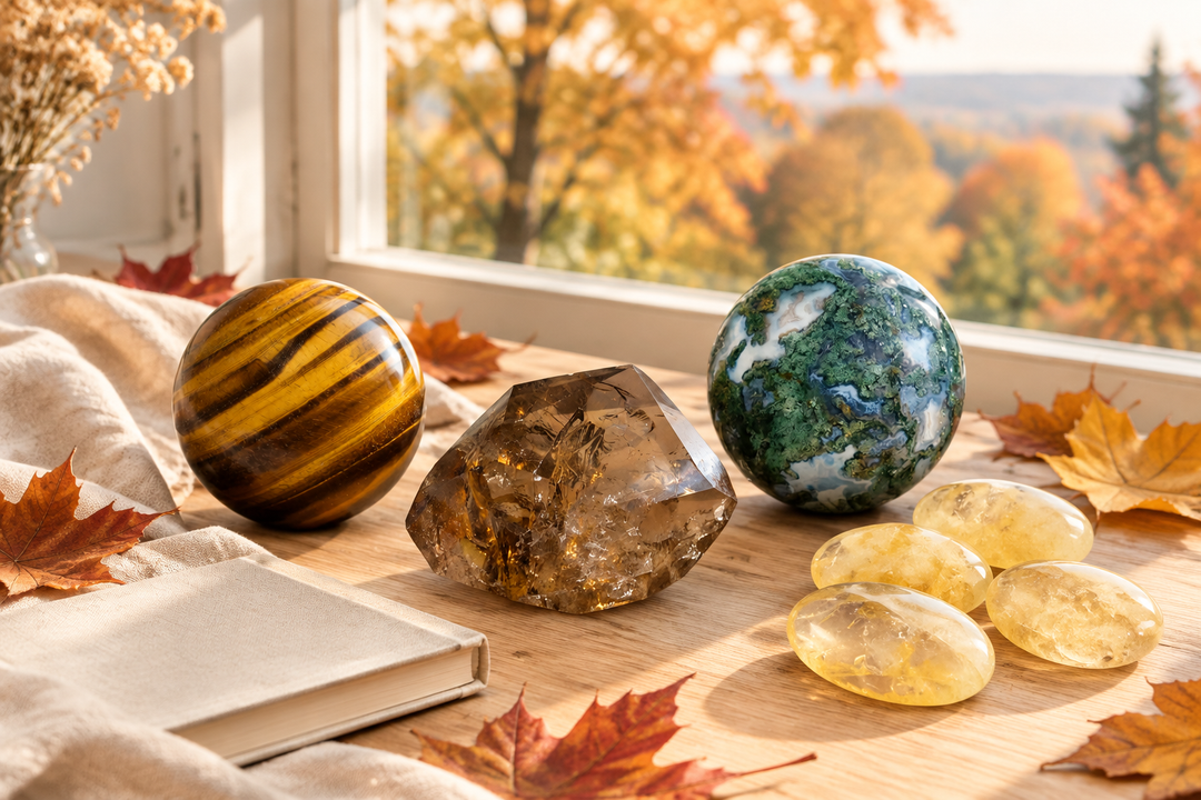 Tiger’s Eye and Moss Agate spheres, Smoky Quartz, and Citrine palm stones on a sunlit wooden table with autumn leaves beside a window.