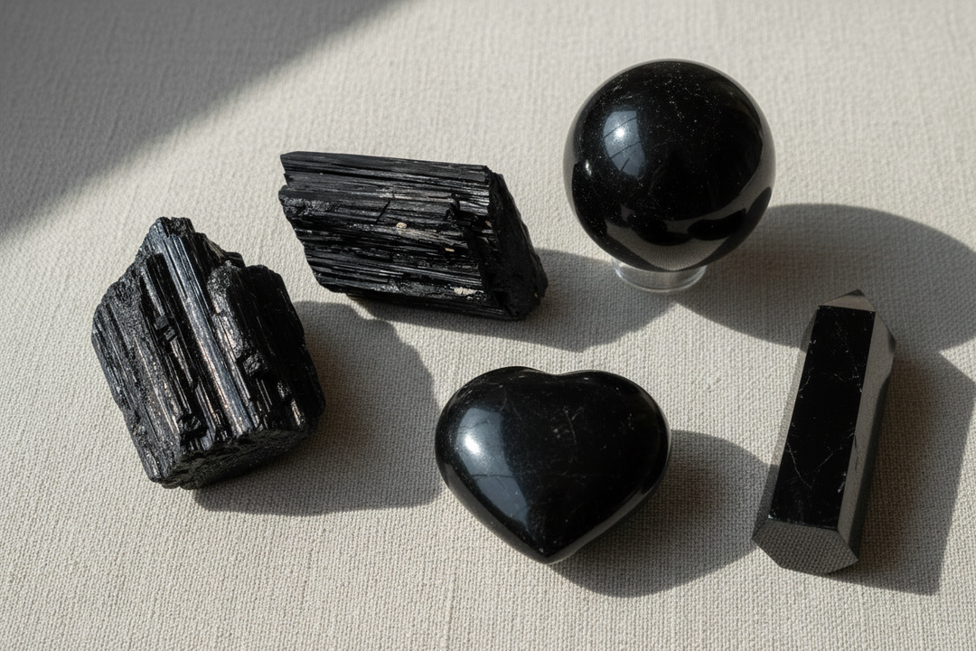 Assortment of black tourmaline crystals: raw chunks, polished sphere, heart, and obelisk on a light surface.