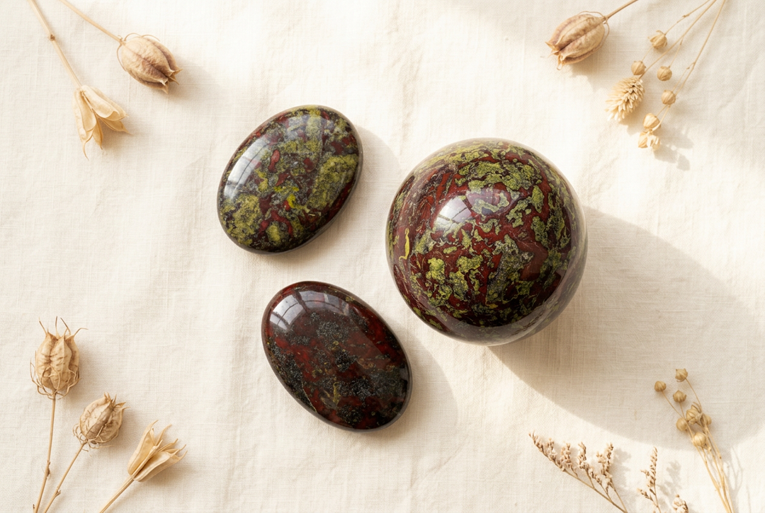 Dragon blood jasper sphere and two polished palm stones with burgundy and olive-green patterns on cream linen with dried botanicals.