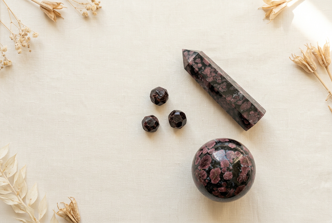 Three rough garnet crystals beside a polished garnet-bearing tower and sphere on warm cream linen with dried botanicals.