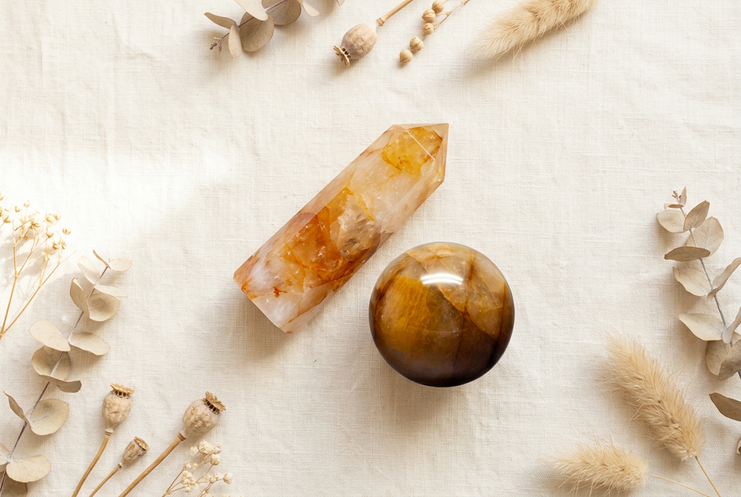Golden healer quartz pointed tower and polished amber-gold sphere on warm cream linen with dried botanicals.