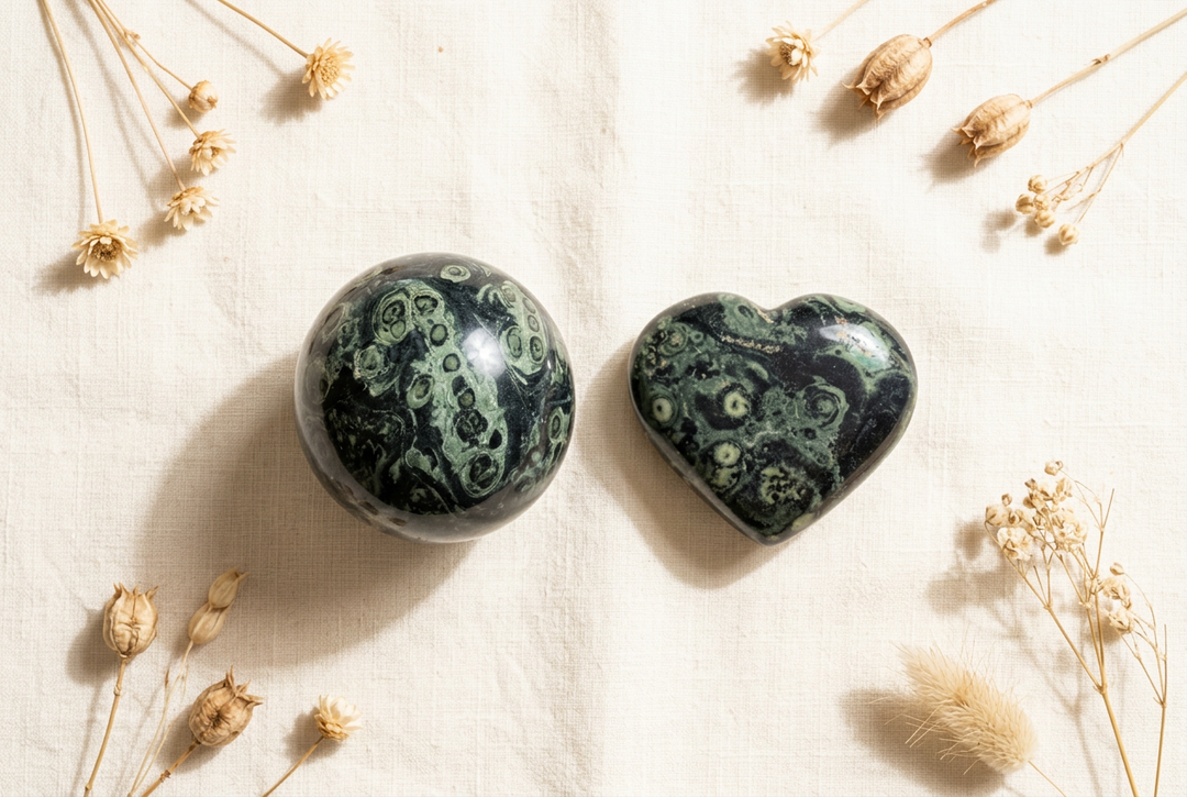 Polished kambaba jasper sphere and heart with sage-green orb patterns on warm cream linen with dried botanicals.
