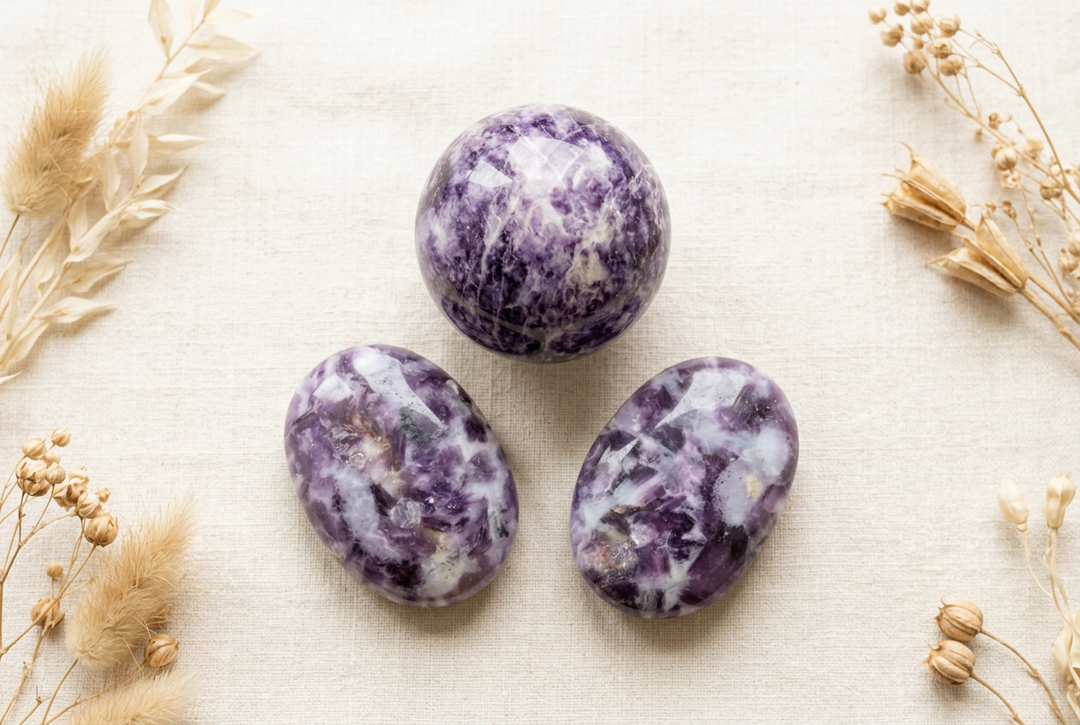 Polished lepidolite sphere and two oval palm stones with purple and white patterns on cream linen with dried botanicals.
