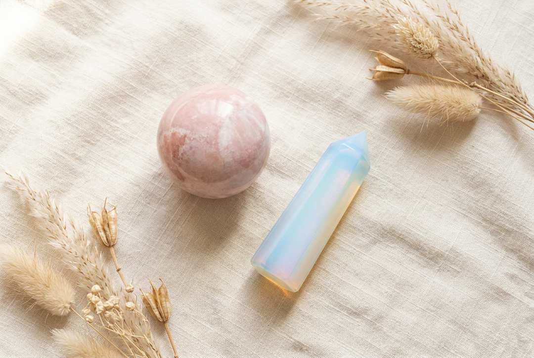 Polished pink opal sphere and milky blue opalite obelisk on warm cream linen with dried botanicals.