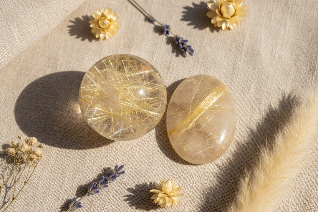Polished rutilated quartz sphere and palm stone with golden needle inclusions on warm linen, soft natural light, spa-inspired flat lay