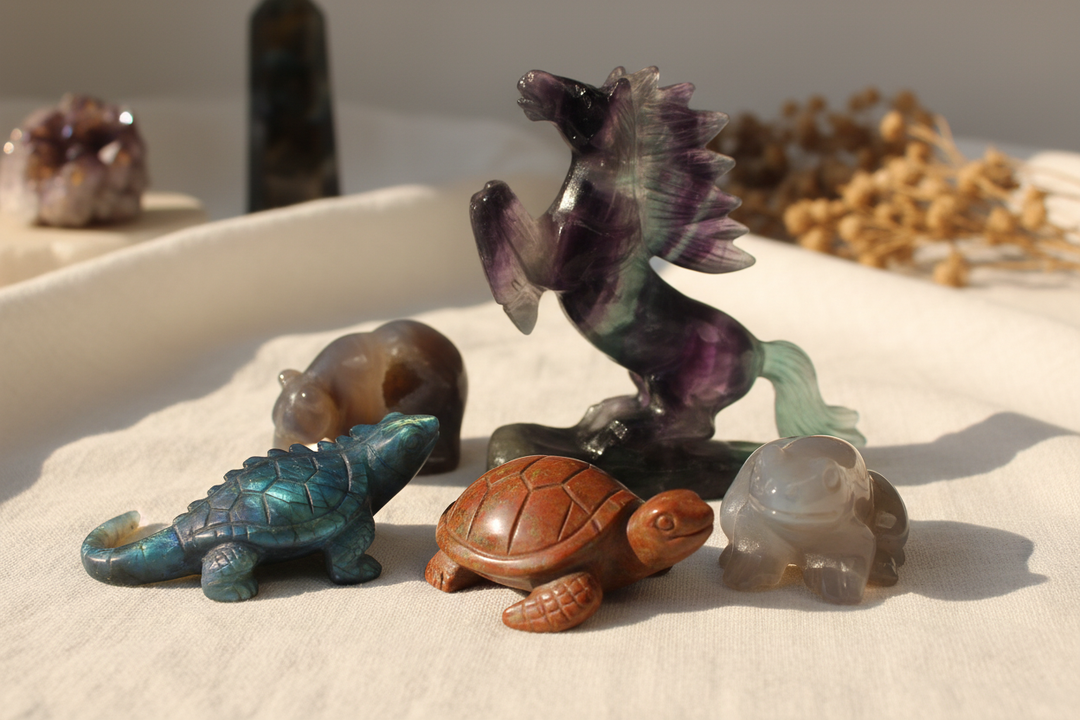 Collection of carved crystal animal figurines including a labradorite lizard, ocean jasper turtle, fluorite horse, agate bear, and crystal frog arranged on white linen