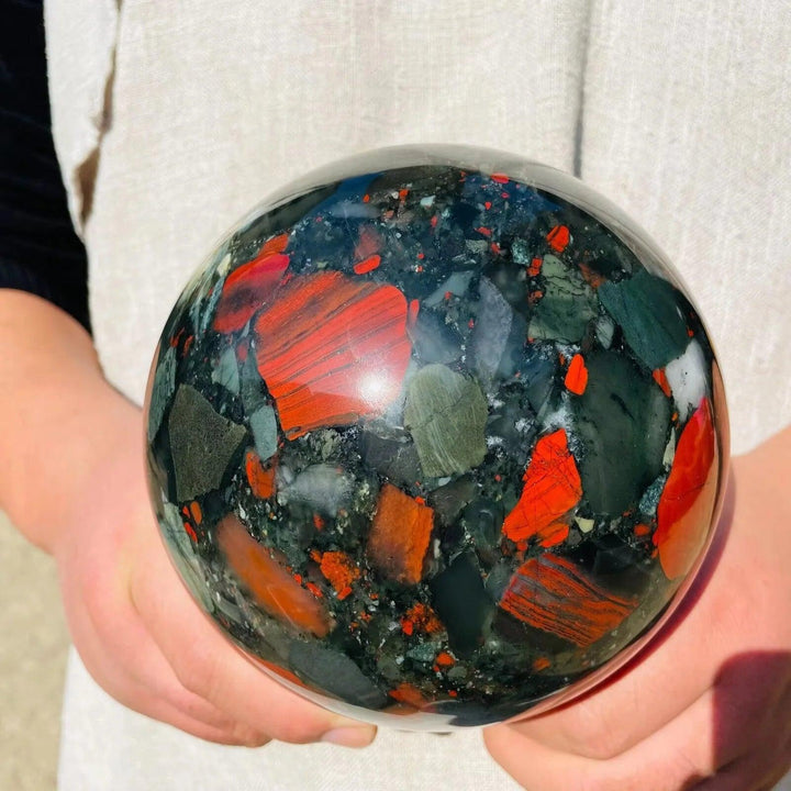 Polished African Bloodstone sphere, 50-85mm, with deep forest green and vivid red iron spots, held in hands.