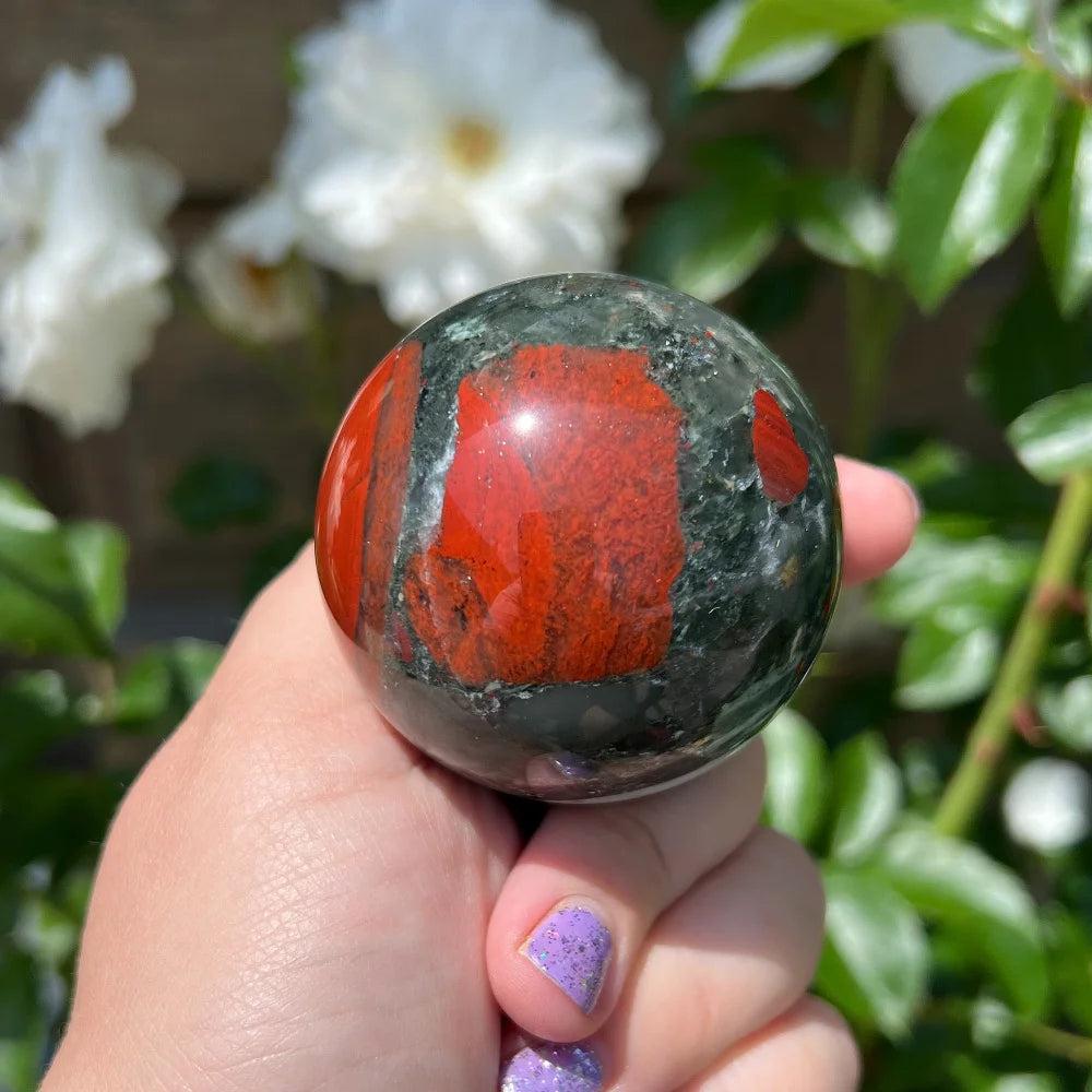 Hand holding a polished African Bloodstone sphere with deep green and vivid red iron spots, against a floral background.