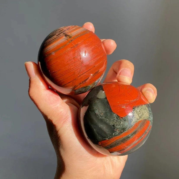 Hand holding two polished African Bloodstone spheres with deep red and dark green patterns for healing and grounding.