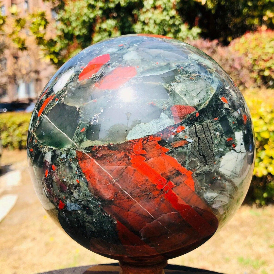 Polished African Bloodstone sphere (50-85mm) with deep forest green and vivid red iron spots, on a wooden stand.