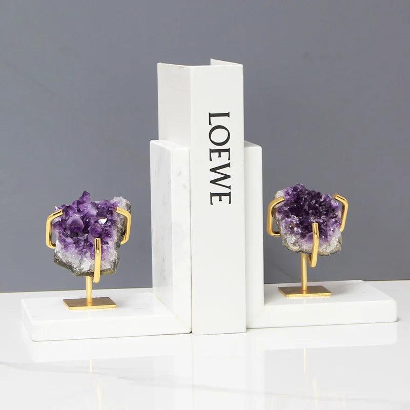 Polished amethyst crystal bookends with gold stands, grounding and stabilizing a white book on a marble base.