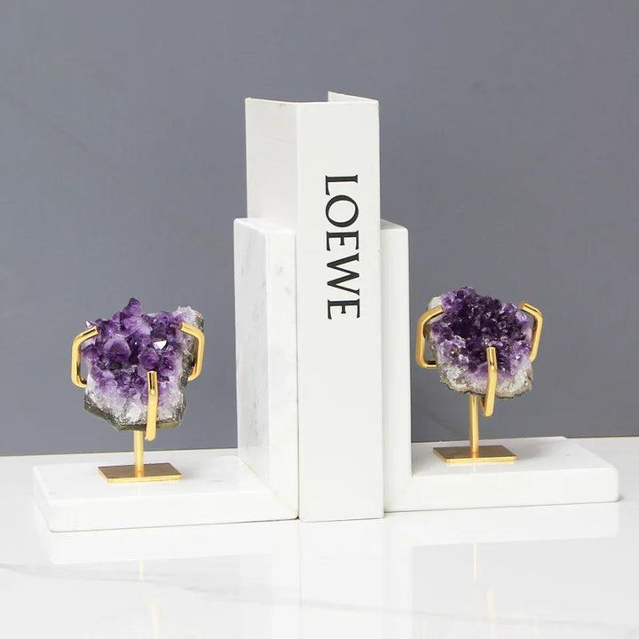 Polished amethyst crystal bookends with gold stands, grounding and stabilizing a white book on a marble base.