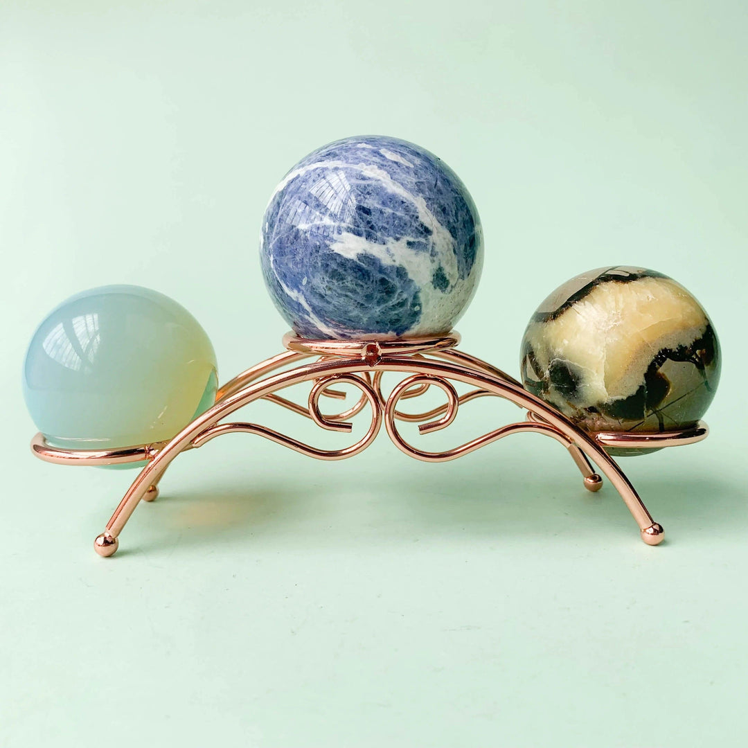 Rose gold arch crystal display stand holding three crystal spheres: blue sodalite, opalite, and jasper.