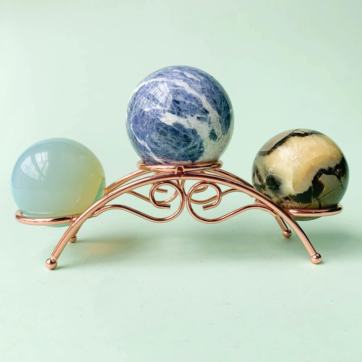 Rose gold arch crystal display stand holding three crystal spheres: blue sodalite, opalite, and jasper.
