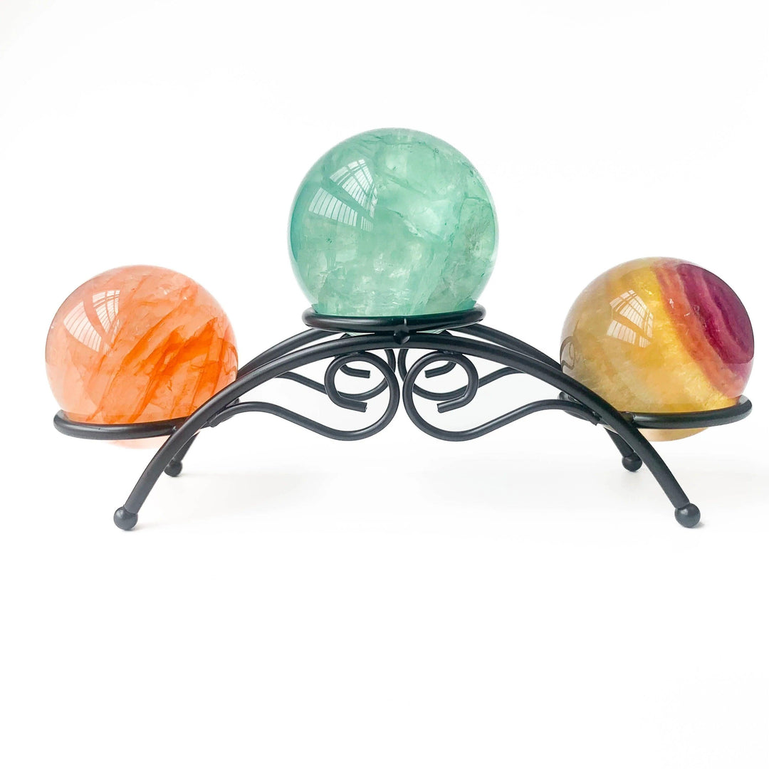 Black arch metal display stand holding three colorful crystal spheres: orange calcite, green fluorite, and rainbow fluorite.