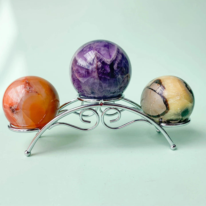 Silvery arch crystal display stand holding three spheres: orange carnelian, purple amethyst, and brown jasper.