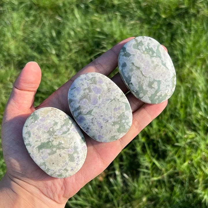 Three polished green and purple Atlantisite Crystal Palm Stones held in a hand for heart chakra healing.