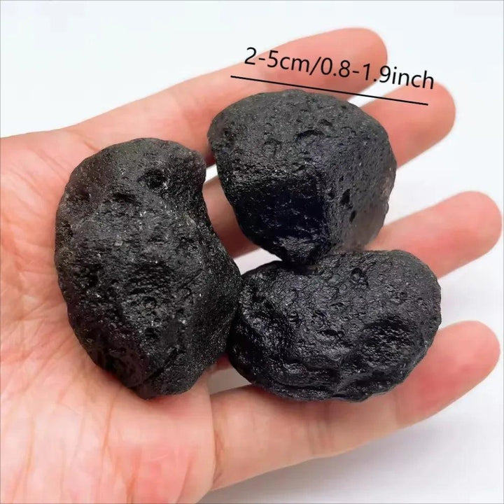 Three rough, dark black Tektite meteorite specimens, 0.8-1.9 inches, in a hand, offering grounding protection.