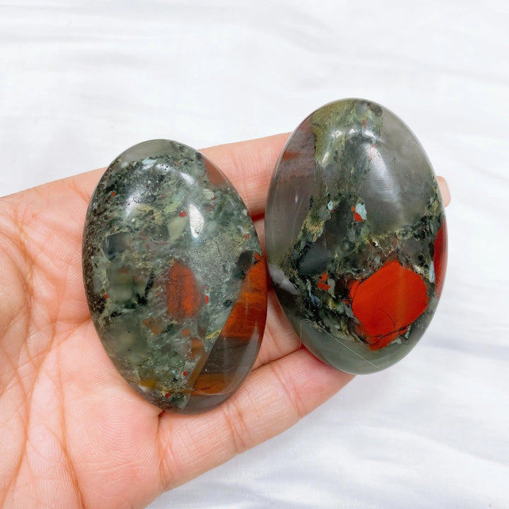 Two polished bloodstone palm stones, dark green with vivid red spots, held in a hand.