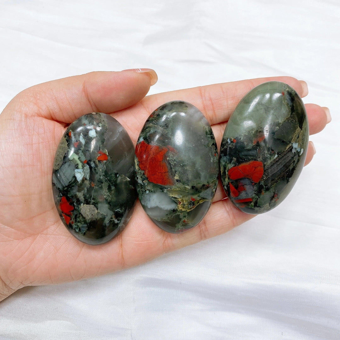 Three polished bloodstone palm stones, dark green with vivid red spots, held in a hand.