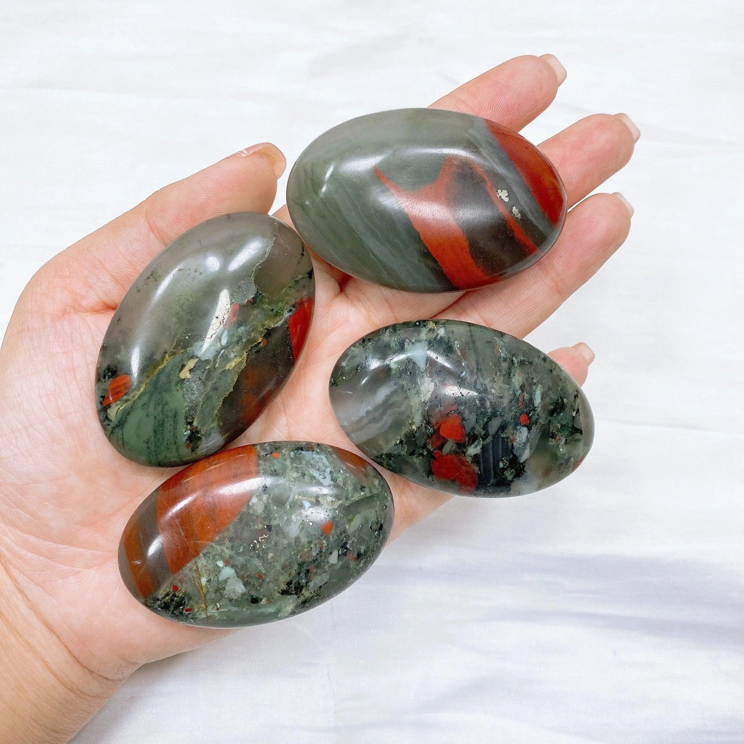Four polished bloodstone palm stones, dark green with vivid red spots, held in an open hand.