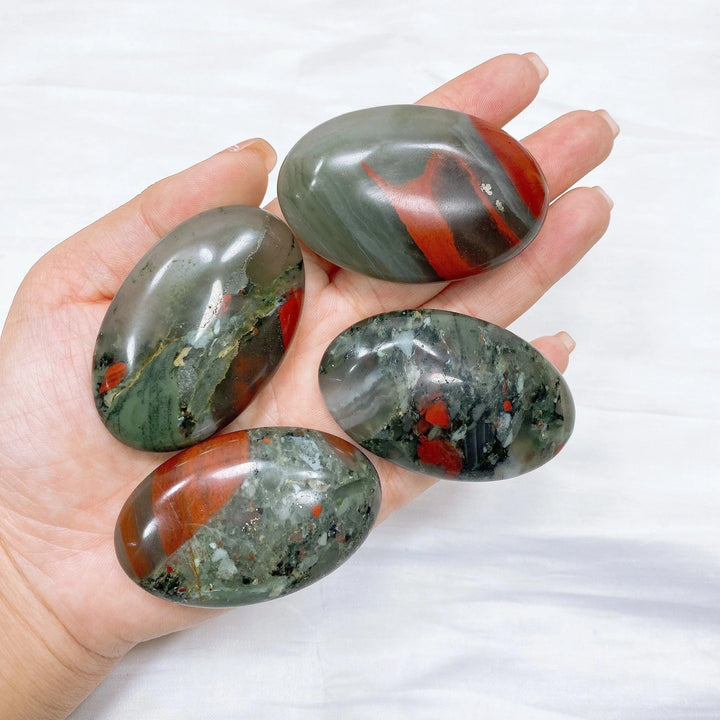 Four polished bloodstone palm stones, dark green with vivid red spots, held in an open hand.