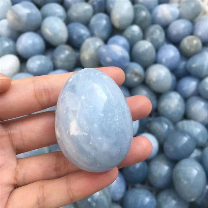 Hand holding a smooth, polished blue Celestite crystal egg, with many similar eggs in the background.
