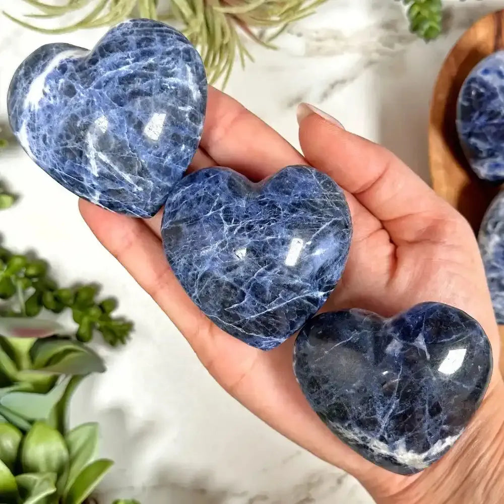 Polished blue sodalite hearts with white veining, held in hand to showcase the Throat Chakra stone's natural beauty.