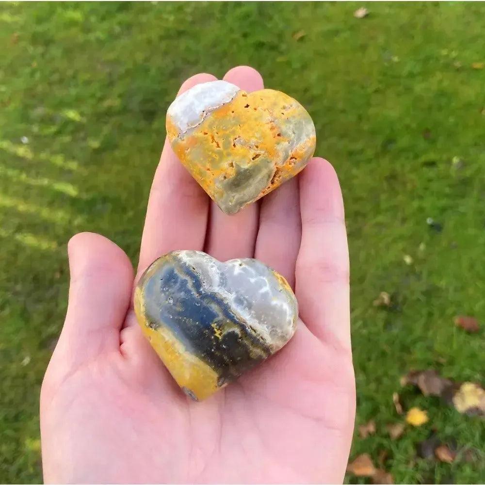 Two polished Bumble Bee Jasper crystal hearts, vibrant yellow, orange, and black, held in a hand.