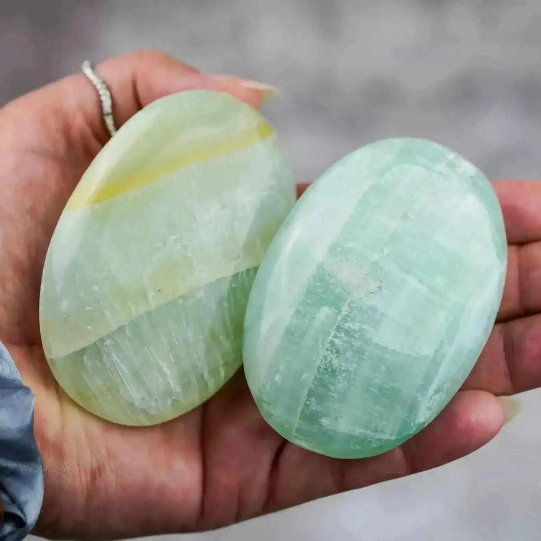 Two smooth, hand-polished Caribbean Calcite palm stones in ocean blue and soft white, held to promote calm and clarity.