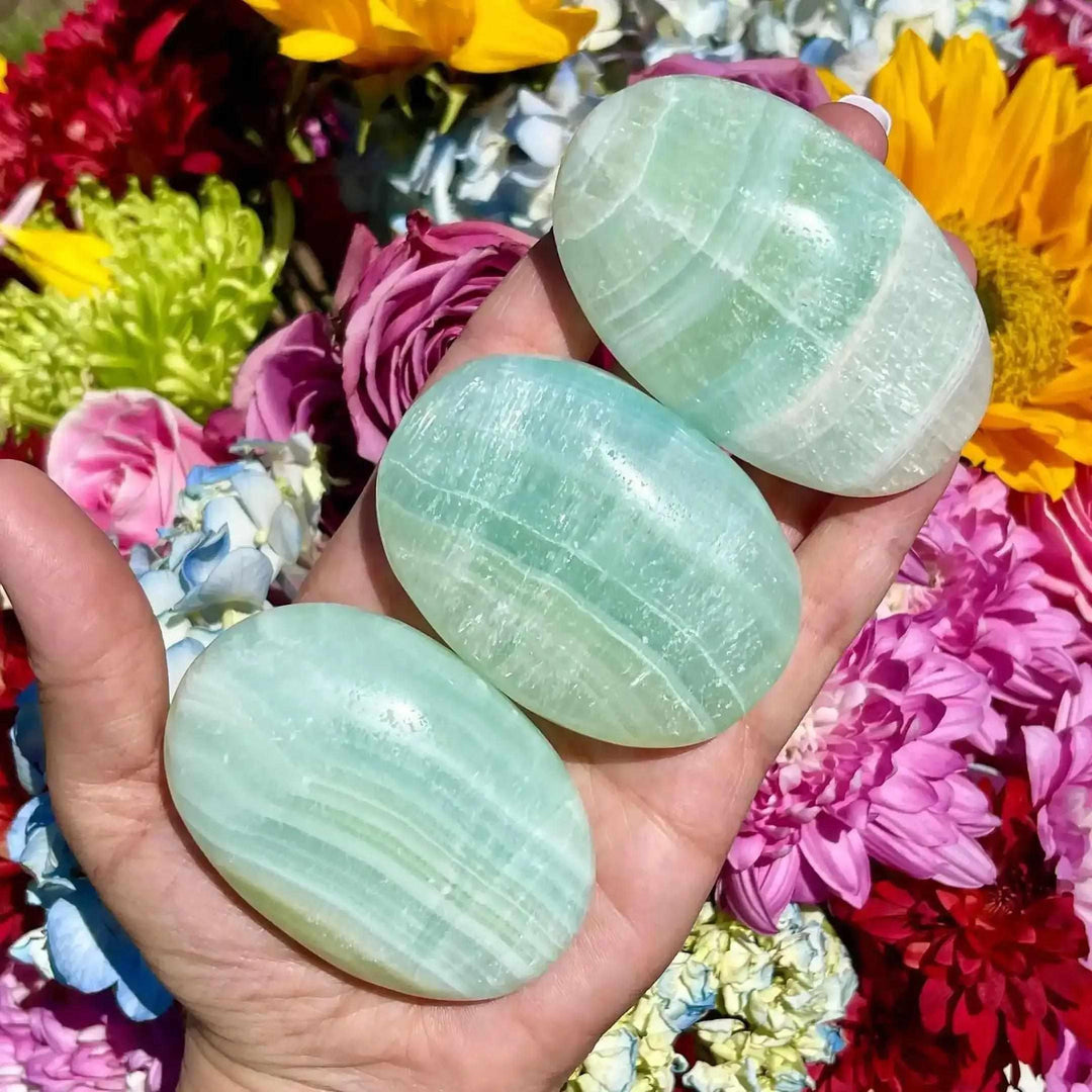 Three smooth, hand-polished Caribbean Calcite palm stones with ocean blue and white swirls held against colorful flowers.