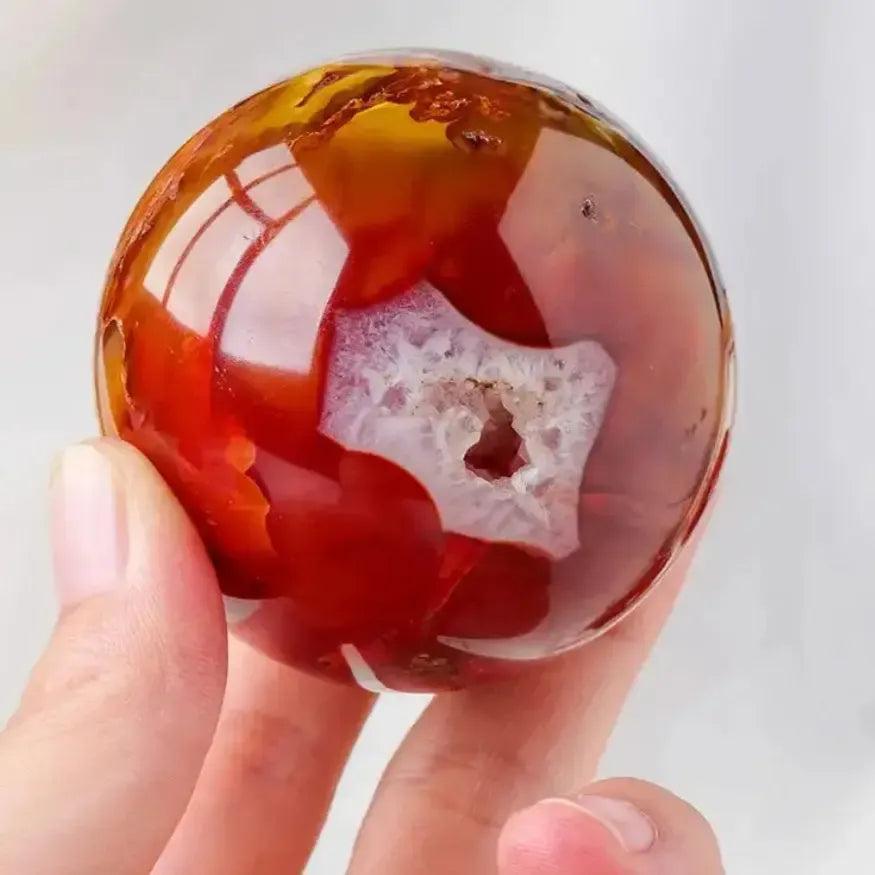 Hand holding a polished, fiery red and orange Carnelian sphere with a sparkling white geode center, radiating desire.