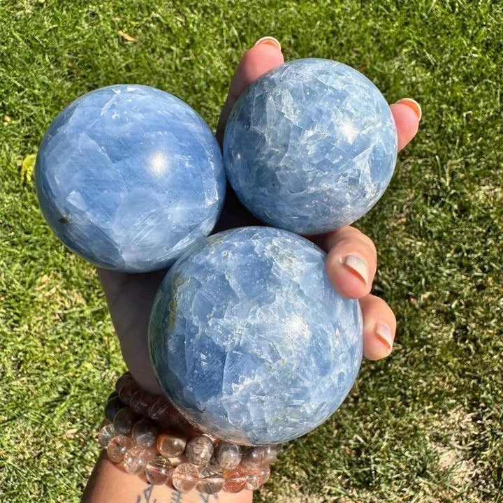 Three polished Celestite crystal spheres in varying soft blue hues, held gently over green grass.