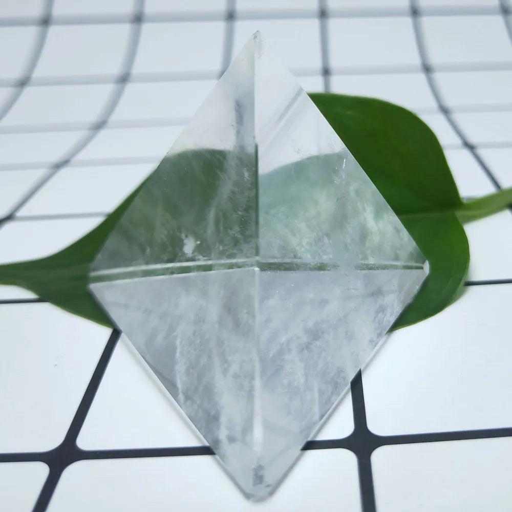 Polished clear quartz crystal tetrahedron pyramid on a white grid with a green leaf, ideal for energy work.