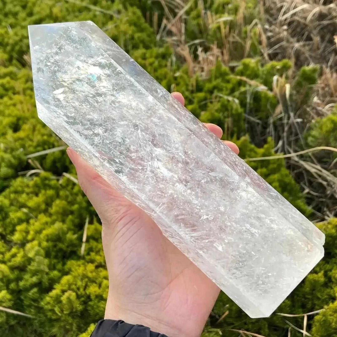 Hand holding a substantial, clear quartz crystal obelisk tower, polished with a single point, against green moss.