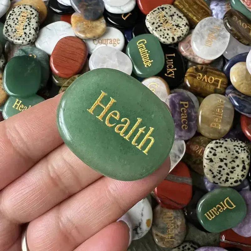 Hand holding a polished green aventurine crystal palm stone engraved with "Health" amidst other colorful inspirational stones.