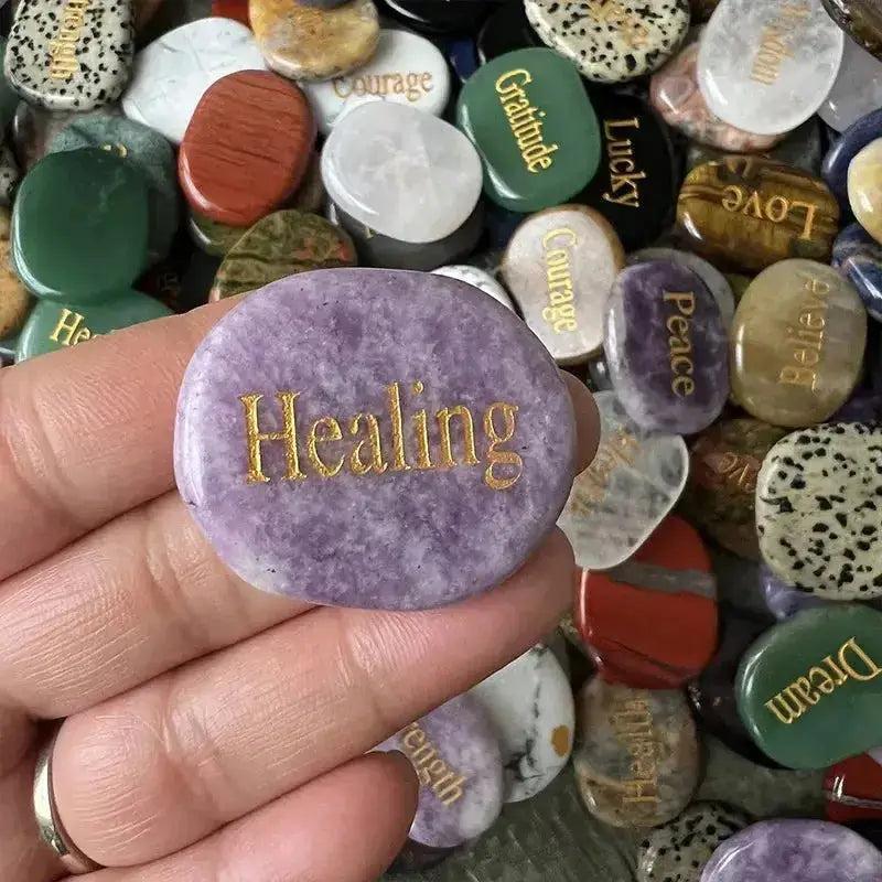 Hand holding a polished purple crystal palm stone engraved with "Healing" in gold, surrounded by assorted inspirational word stones.