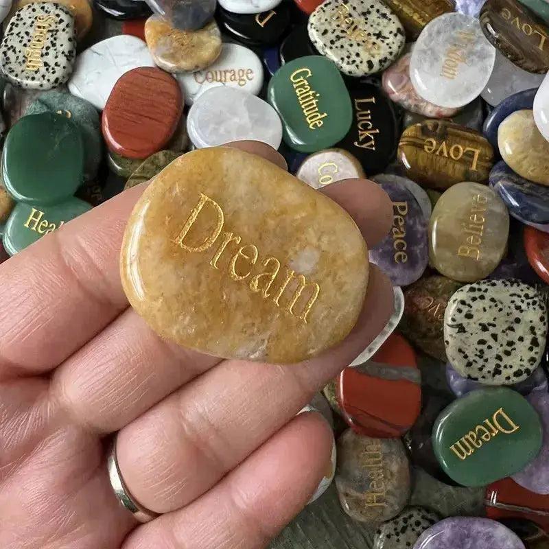 Hand holding a polished yellow crystal palm stone engraved with "Dream," surrounded by other colorful inspirational stones.