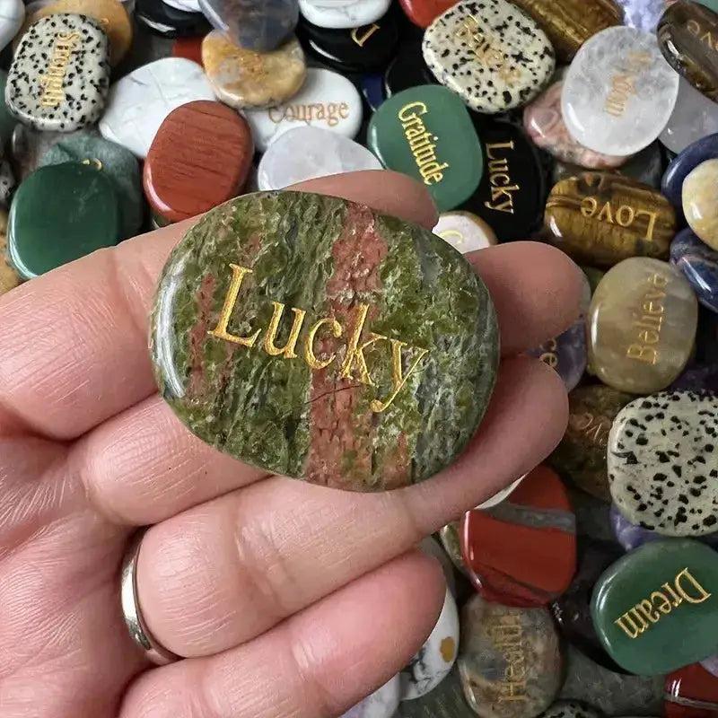Hand holding a polished green and red crystal palm stone engraved with "Lucky," surrounded by other inspirational stones.