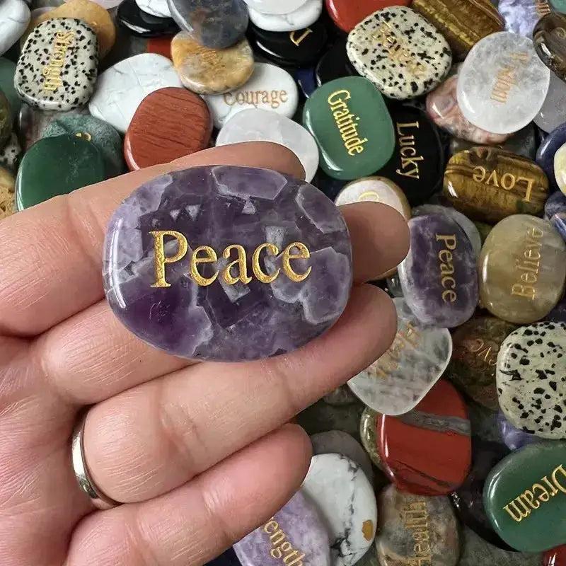 Hand holding a polished amethyst palm stone engraved with "Peace" among assorted inspirational crystal stones.