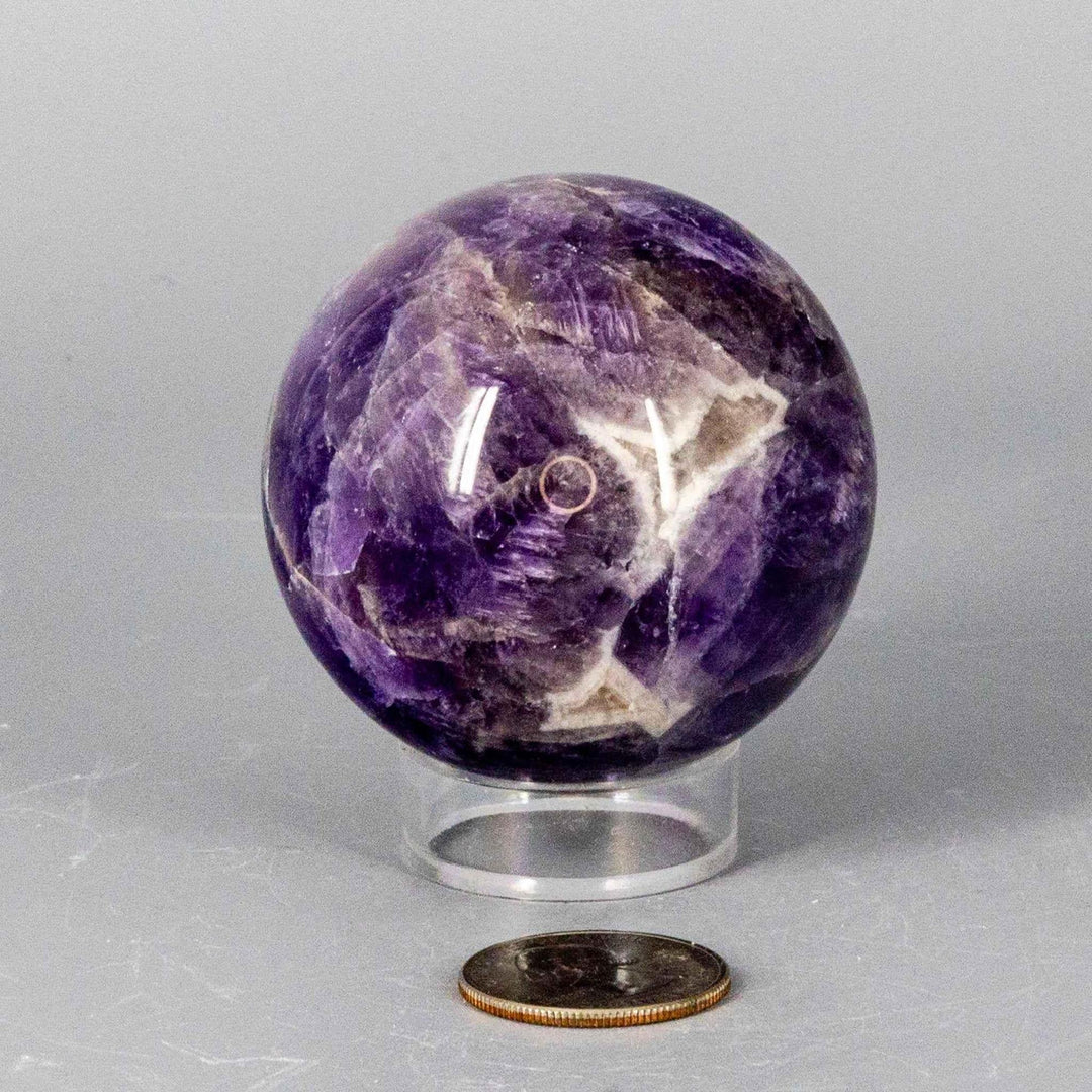 Dream Amethyst Sphere 51-57mm: A polished purple and white banded crystal sphere on a clear stand, next to a quarter.