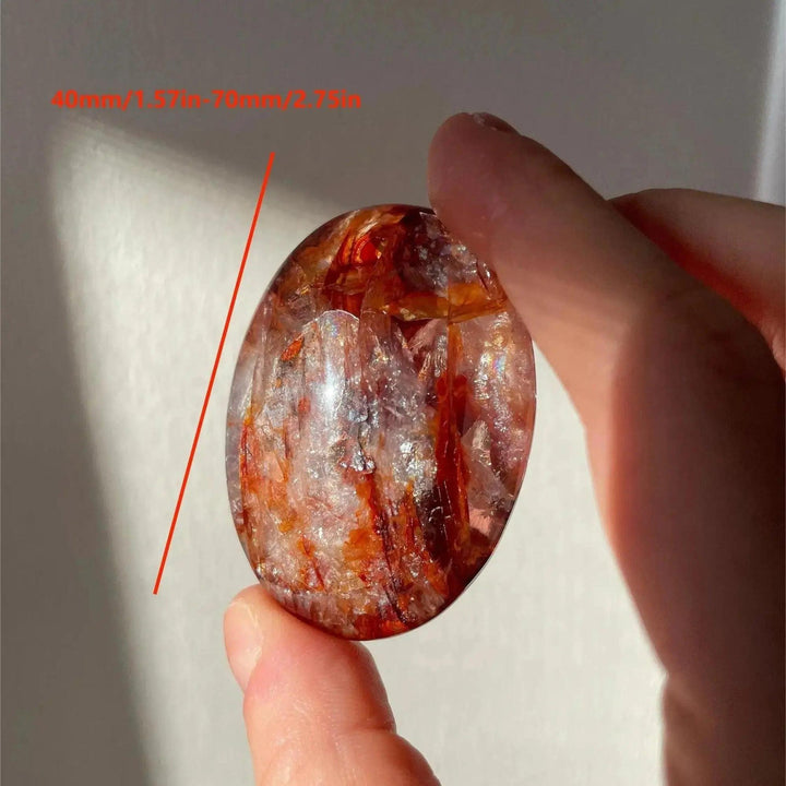 Crystal Destiny Fire Quartz palm stone, clear with fiery red hematite, smooth oval, held between fingers.