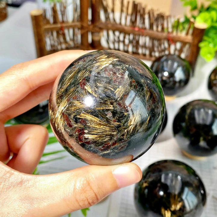 Hand holding a polished black crystal sphere with golden fibrous inclusions, from Crystal Destiny.
