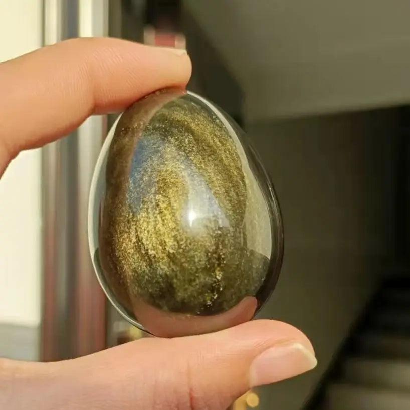 Hand holding a polished Gold Sheen Obsidian crystal egg, showcasing its dark volcanic glass with golden shimmer.