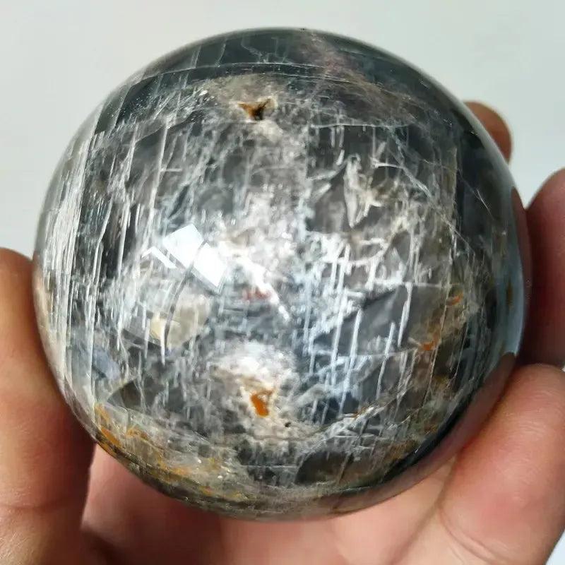 Hand holding a dark, shimmering black obsidian crystal sphere with white and silver inclusions for purification and cleansing.