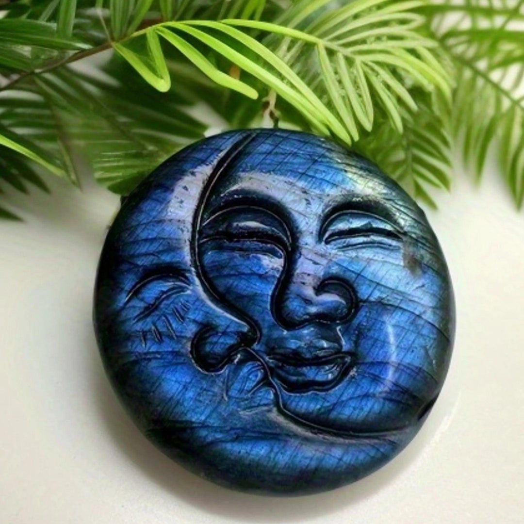 Shimmering blue labradorite crystal moon and sun carving, symbolizing intuition and clarity, against green foliage.