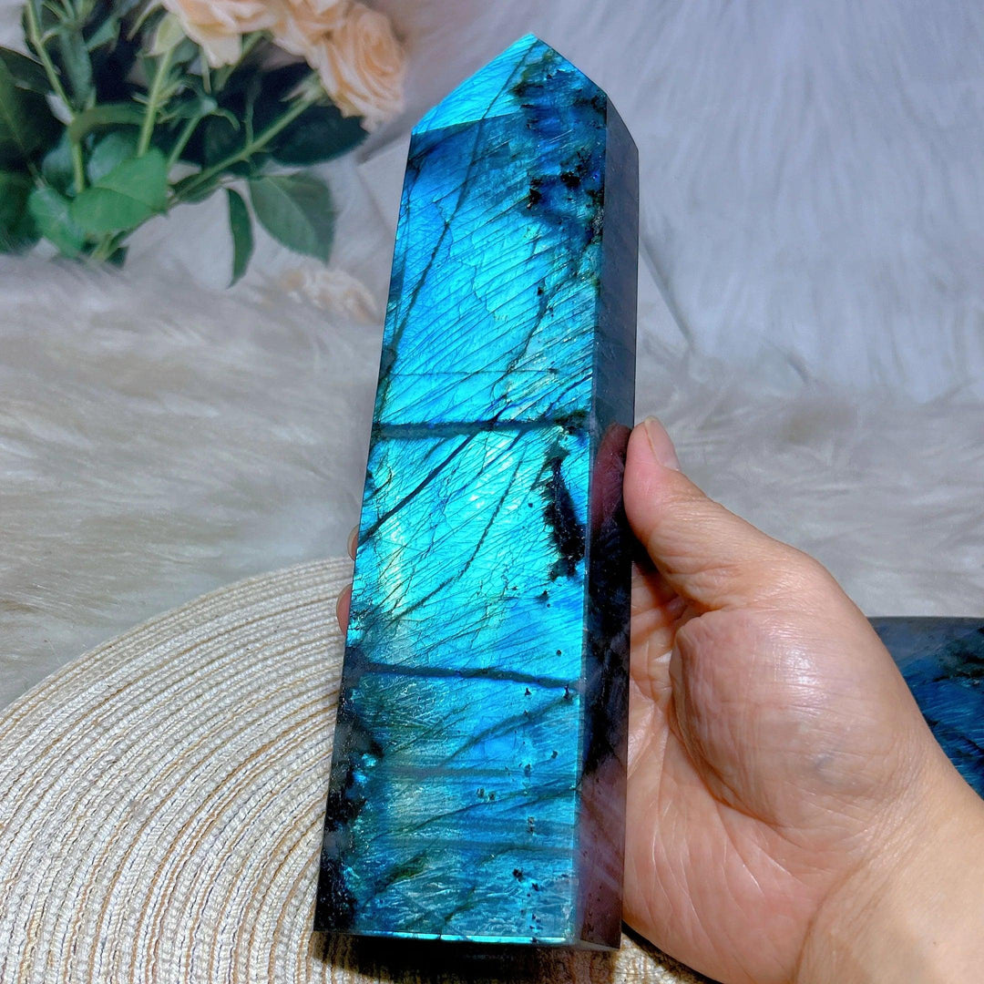 Hand holding a tall, polished Labradorite crystal tower with vibrant blue and teal labradorescence.