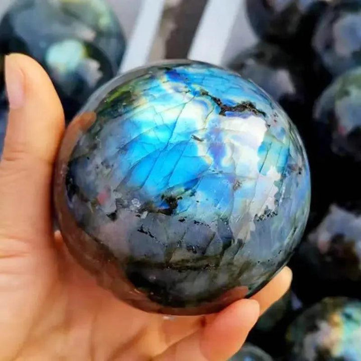 Hand holding a polished Labradorite sphere with vibrant blue, green, and gold iridescent flashes from Crystal Destiny.