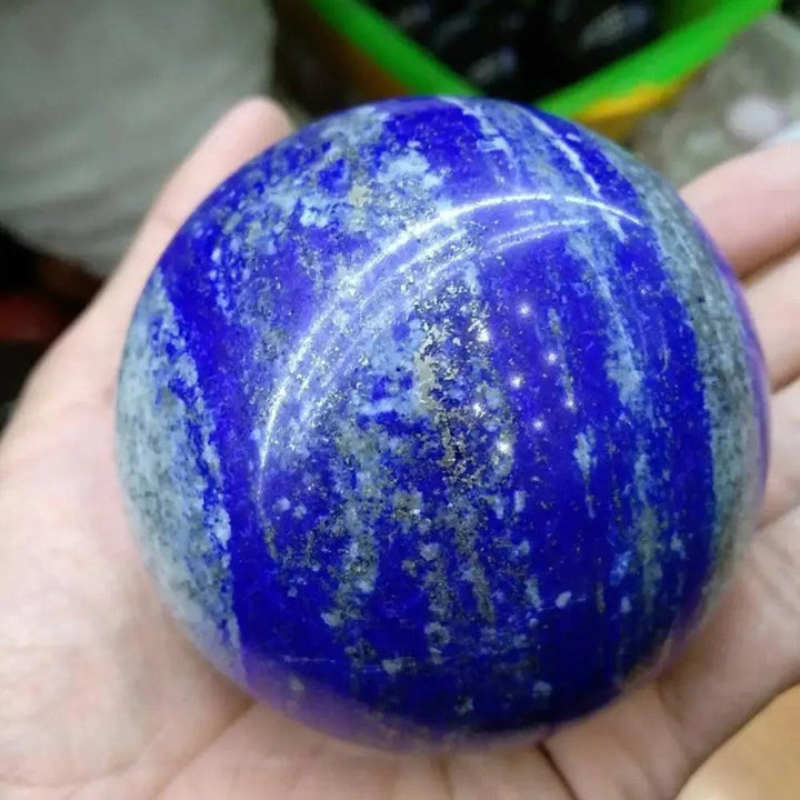 Polished Lapis Lazuli Crystal Sphere, deep celestial blue with golden pyrite flecks and white calcite veining, held in a hand.