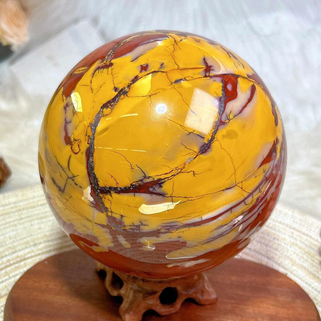Large 2695g Mookaite jasper sphere with red, yellow, and cream swirls on a wooden stand for grounding energy.